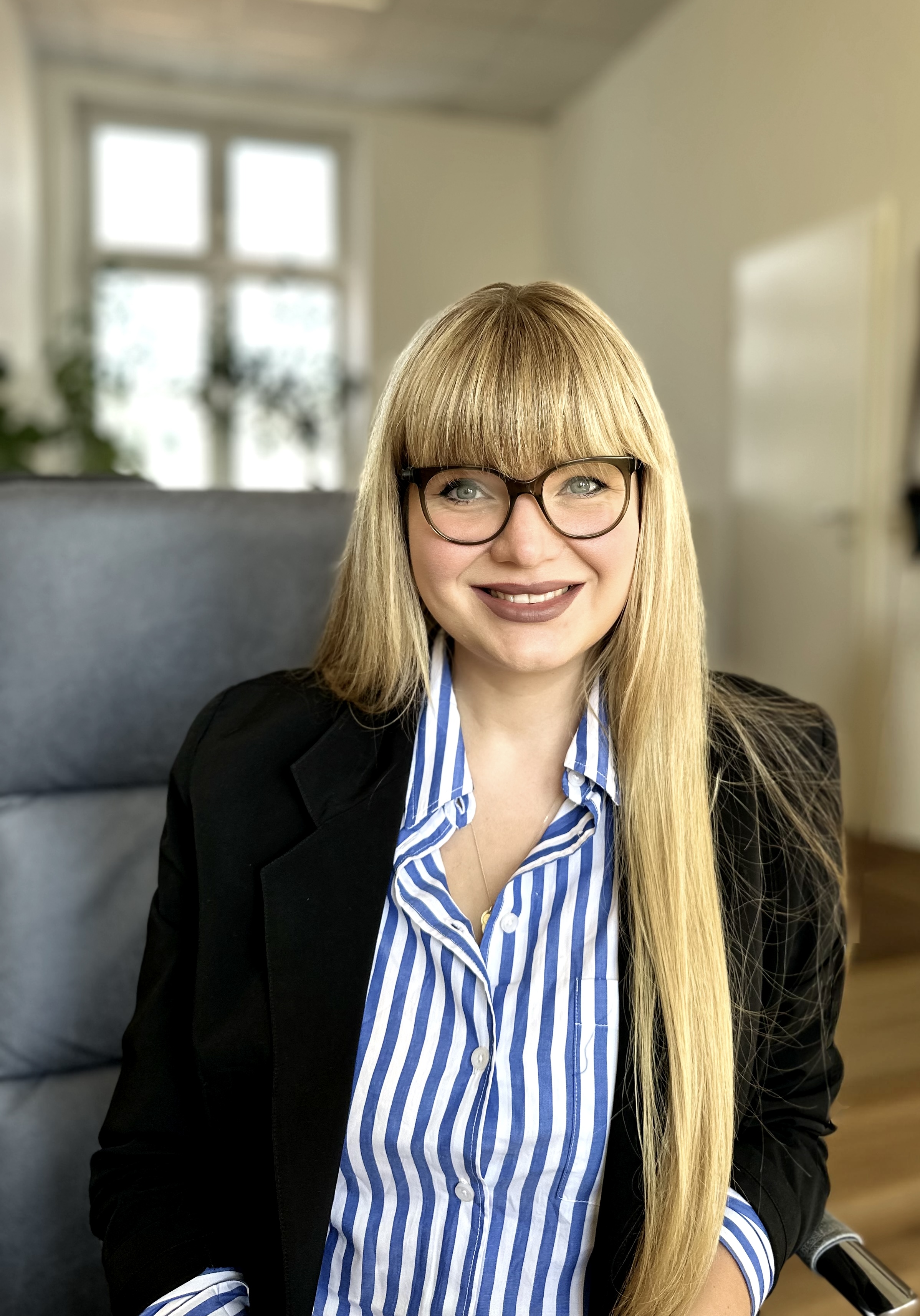 Person mit langen blonden Haaren sitzt in einem hellen Büro; schwarzer Blazer, blau‑weiß gestreiftes Hemd.