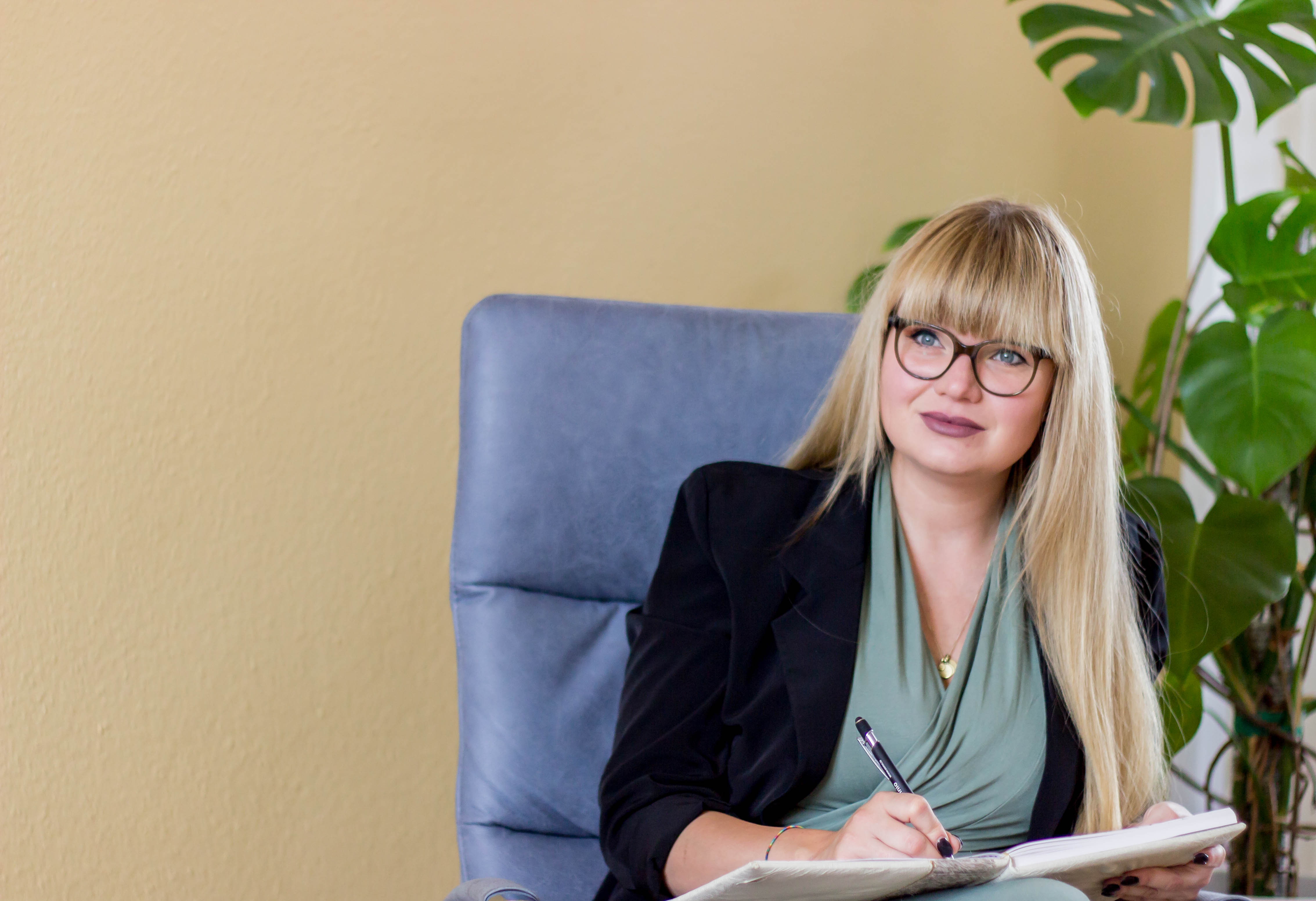 Eine Frau mit langen blonden Haaren und Brille sitzt in einem Sessel, schreibt in ein Notizbuch und wirkt aufmerksam und freundlich. Im Hintergrund ist eine beige Wand mit einer grünen Pflanze zu sehen.